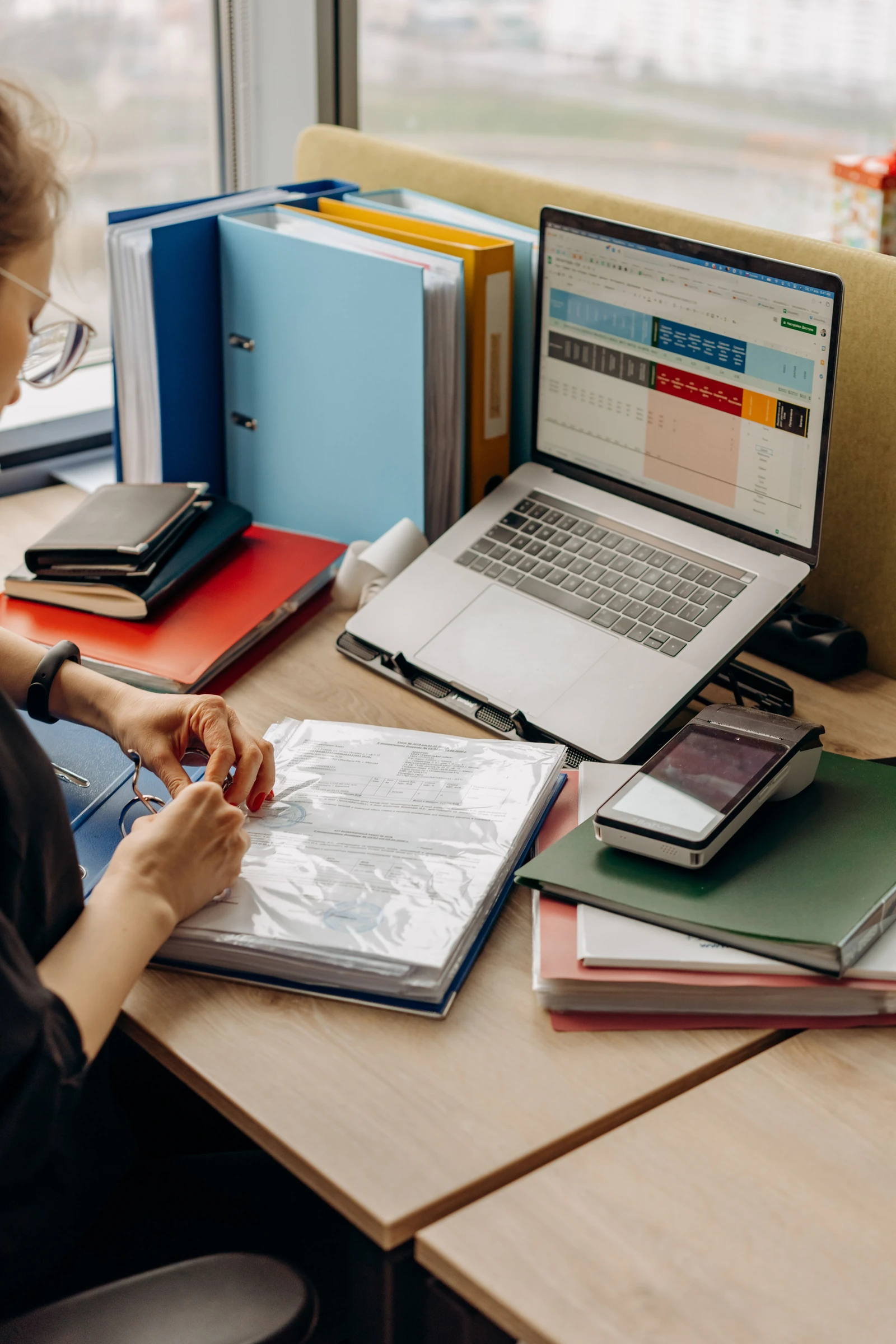Supplied photograph of a person reviewing paperwork beside a laptop, colourful binders and a payment terminal