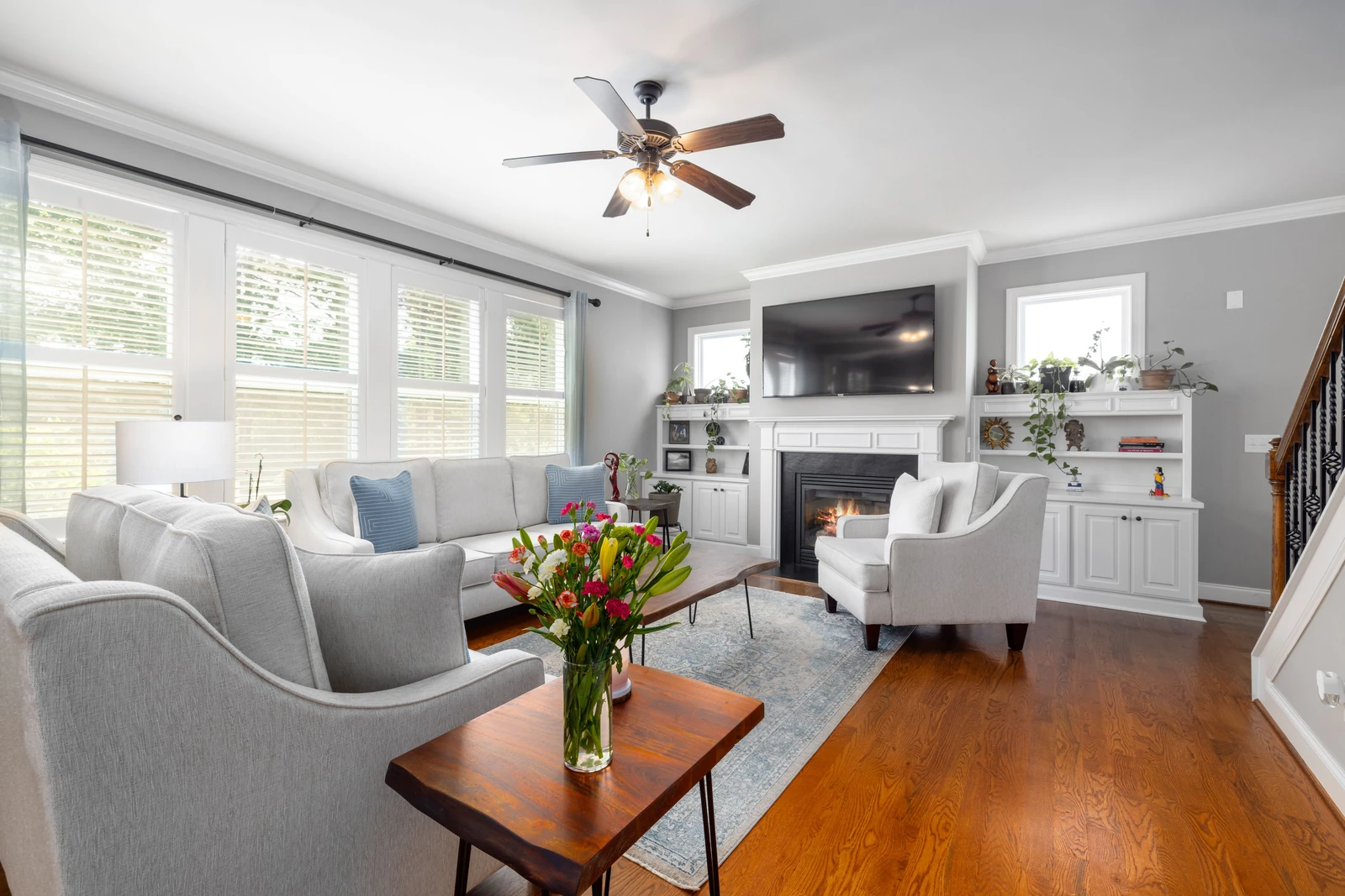 Supplied property photograph of a bright living room with pale seating, a fireplace, ceiling fan and flowers on timber tables