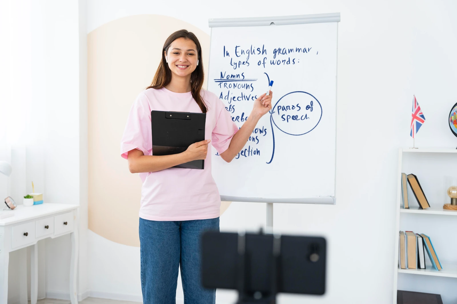 Supplied photograph of a woman in a pink shirt teaching English grammar beside a whiteboard and a recording phone